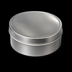 Puede incluir: Una pequeña lata redonda de color plateado con tapa. La lata es de metal y tiene un acabado liso y mate. La tapa encaja perfectamente en la parte superior de la base. El fondo es negro liso.