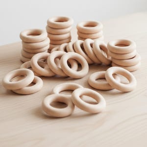 May include: A collection of 50 unfinished wooden craft rings, perfect for DIY projects. The rings are stacked and scattered on a light-colored wooden surface. The natural wood color is visible, and the rings are smooth and round.