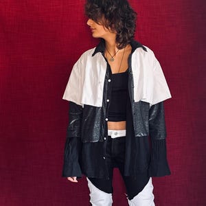 May include: A long-sleeve button-down shirt featuring a white upper section and a black lower section with a subtle metallic sheen. The shirt is worn over a black tank top and paired with black and white color-blocked pants with a white belt.