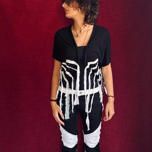May include: A black short-sleeved top featuring white frayed fabric strips hanging from the front, paired with black pants that have large white fabric panels sewn into the legs and a white waistband, creating a high-contrast monochromatic look.
