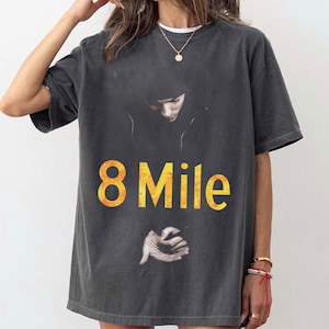 May include: A charcoal gray short-sleeve t-shirt featuring a graphic print of a person in a hooded jacket with the text 8 Mile in large yellow letters and an image of hands below the text, printed on the front of the garment.
