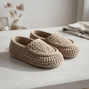 May include: A pair of beige slip-on slippers crafted from thick, chunky yarn in a crochet knit pattern. The slippers feature a rounded toe design with a raised, stitched seam detail along the top edge and a solid, textured construction throughout.