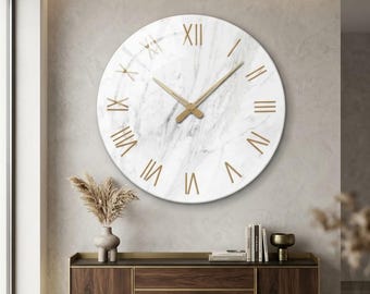 May include: A large circular wall clock featuring a white marble-patterned face with gold-colored Roman numerals and matching gold-colored hour and minute hands. The clock has a smooth, reflective surface and a clean, simple design.