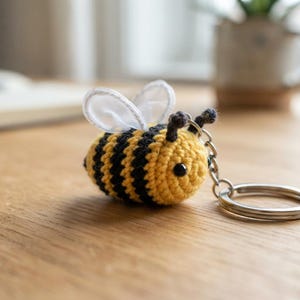 Könnte beinhalten: Ein kleiner gehäkelter Bienen-Schlüsselanhänger mit einem runden Körper mit gelben und schwarzen Querstreifen, zwei kleinen schwarzen Fühlern und durchscheinenden weißen Stoffflügeln auf der Oberseite, befestigt an einem runden silbernen Metallring.