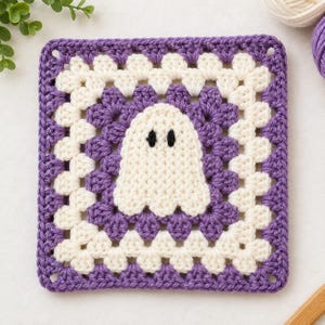 May include: A square crochet granny square featuring a central white ghost motif with two black embroidered eyes. The square is constructed with alternating rounds of purple and cream-coloured yarn in a classic cluster stitch pattern.
