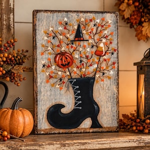 May include: A rectangular wooden sign with a distressed light gray finish featuring a painted black boot. Emerging from the boot are branches with orange, yellow, and white leaves, a jack-o'-lantern, a witch hat, and a piece of candy corn.