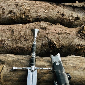 May include: A silver-coloured metal sword with a black wrapped handle and a flared pommel, positioned next to a matching black scabbard with silver metal accents. The sword features a straight crossguard and a polished blade surface.