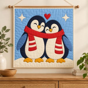May include: A square quilted wall hanging featuring two penguins with black, white, and yellow bodies, wearing a shared long red scarf with white stripes. A red heart sits above them against a light blue background with two white stars.