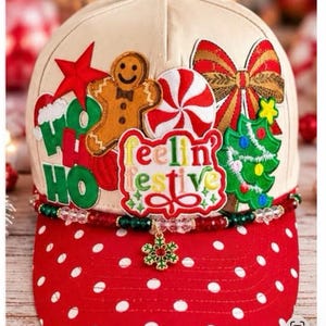 May include: A cream-colored baseball cap with a red polka-dot brim. The front features patches of a gingerbread man, a red star, a peppermint candy, a bow, and a Christmas tree. Text reads "feelin festive" and "HO HO HO". A beaded trim with a snowflake charm.