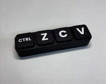 May include: A black rectangular macro keypad featuring four square keys arranged in a horizontal row. Each key has a textured surface and displays a white letter or label: CTRL, Z, C, and V, representing common computer shortcut commands.