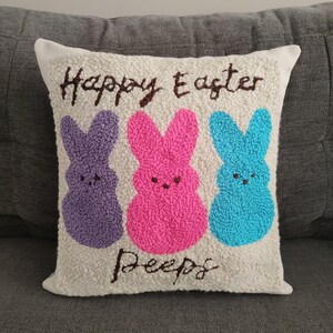 Puede incluir: Una almohada blanca con las palabras "Happy Easter Peeps" bordadas en hilo marrón. Tres Peeps coloridos están bordados en el centro de la almohada: morado, rosa y azul.