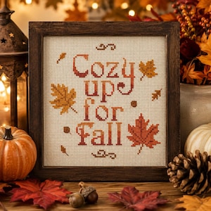 May include: A square cross-stitch embroidery in a dark brown wooden frame featuring the text "Cozy up for Fall" in orange and brown thread, surrounded by stitched autumn leaf motifs on a cream-coloured fabric background.