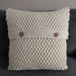 Puede incluir: Una almohada de punto de ganchillo de color crema con un patrón texturizado y dos botones de madera. La almohada está sentada en un sofá gris.