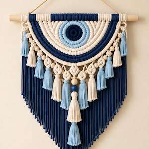 May include: A macrame wall hanging featuring a concentric eye pattern in navy blue, light blue, and cream cotton cord. The design includes wooden beads, multiple hanging tassels in matching colors, and a wooden dowel with a hanging rope.