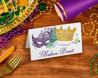 May include: A white card with a purple and teal mask and a gold crown design. The card reads "Madison Benoit" in purple script. The image also includes a gold tray, beads, and a purple party horn.