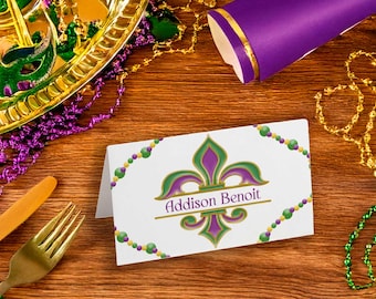 May include: A white place card with a gold and green fleur-de-lis design. The name "Addison Benoit" is printed in black lettering on the card.