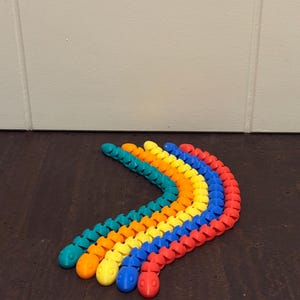 Könnte beinhalten: Fünf bewegliche, segmentierte Kunststoffspielzeuge in Form von Schlangen oder Raupen, die in einer Kurve angeordnet sind. Die Spielzeuge sind in den Farben Türkis, Orange, Gelb, Blau und Rot gehalten und haben jeweils einen runden Kopf mit zwei kleinen Vertiefungen als Augen.