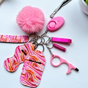 Pode incluir: Um conjunto de porta-chaves marmoreado em rosa e laranja com um pompom rosa felpudo, um alarme de segurança pessoal rosa, um abre-garrafas rosa, um pequeno cilindro rosa, uma ferramenta abre-portas rosa, uma alça e dois suportes de tecido.