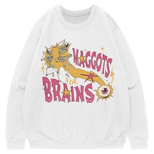May include: A white crewneck sweatshirt featuring a graphic print of two skeletons dancing, a large eyeball, and the text "MAGGOTS FOR BRAINS" in pink letters. The design includes yellow star accents and a yellow trail connecting the eye to the skeletons.