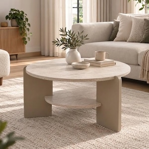 May include: A round coffee table with a light beige stone-textured top and a lower shelf, supported by two thick, curved, solid beige legs. The table surface holds a small white vase with green foliage, a small bowl, and a mug on books.