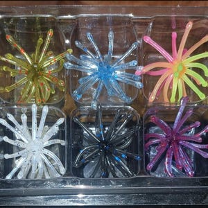 May include: A set of six multi-colored fishing lures, each featuring a central body with numerous radiating flexible tentacles. The lures are arranged in a clear plastic tray with individual compartments, displaying colors including olive, blue, pink, white, black, and purple.