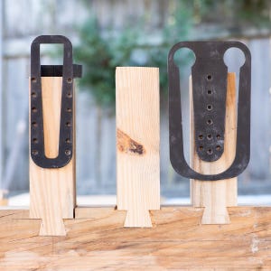 Op de afbeelding: Drie houten planken met zwaluwstaartverbindingen, elk met een andere zwarte metalen beugel. De beugels hebben verschillende vormen en gatenpatronen. Het hout is licht van kleur en het metaal is donker.