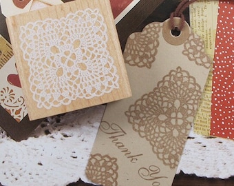 May include: A wooden stamp featuring a white lace design and a tag with a brown lace pattern and the words "Thank You". The stamp and tag are set against a backdrop of assorted paper scraps, including red dotted paper.