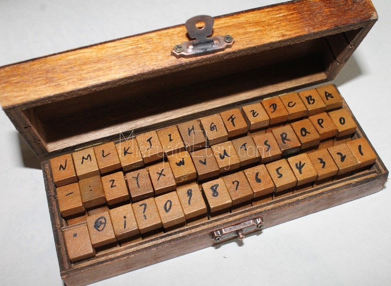 Pu&ograve; includere: Una scatola di legno con un coperchio incernierato che contiene un set di timbri con lettere e numeri. I timbri sono disposti in file e sono in legno con inchiostro nero.