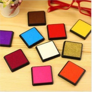 May include: A collection of colorful square ink pads in various shades, including red, blue, yellow, and pink. Each pad has a black border and sits on a light wooden surface. A few small flowers and red glasses are in the background.