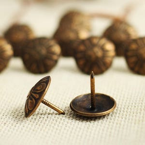 Puede incluir: Primer plano de dos tachuelas de lat&oacute;n antiguo con cabezas decorativas. Las tachuelas tienen un clavo puntiagudo y una parte superior redonda y adornada. Las tachuelas est&aacute;n sobre una superficie de tela marr&oacute;n claro.