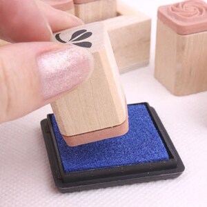 May include: A hand presses a wooden stamp onto a blue ink pad. Several other stamps with floral designs are visible. The stamps have light brown wooden blocks and pink rubber stamp surfaces. The ink pad is black.