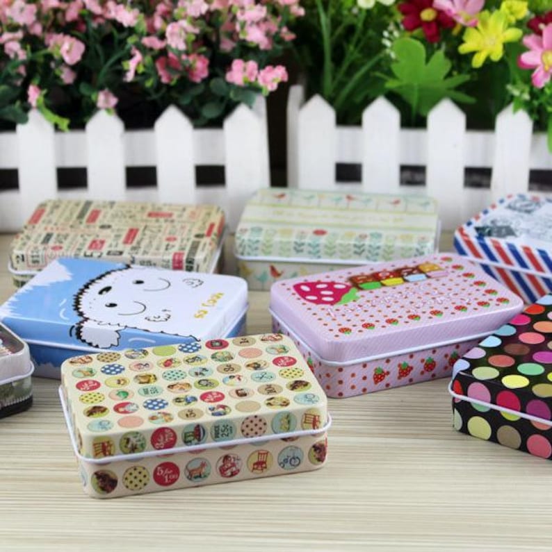 May include: Assortment of small, rectangular, metal tins with various colorful patterns. The tins feature designs of polka dots, strawberries, a dog, and a floral pattern.