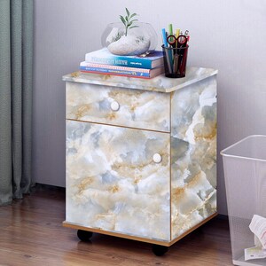 May include: A two-drawer rolling cabinet with a marble-patterned finish. The cabinet has two white knobs and is topped with a stack of books and a glass vase with a small plant.