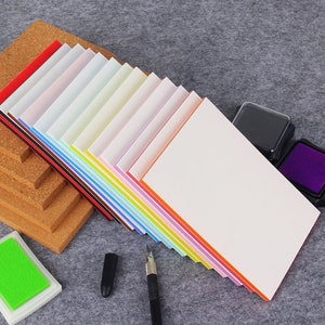 May include: A stack of colorful paper with a white top sheet, two ink pads, a pen, and a cork board. The paper has a variety of colors, including red, orange, yellow, green, blue, purple, and pink.