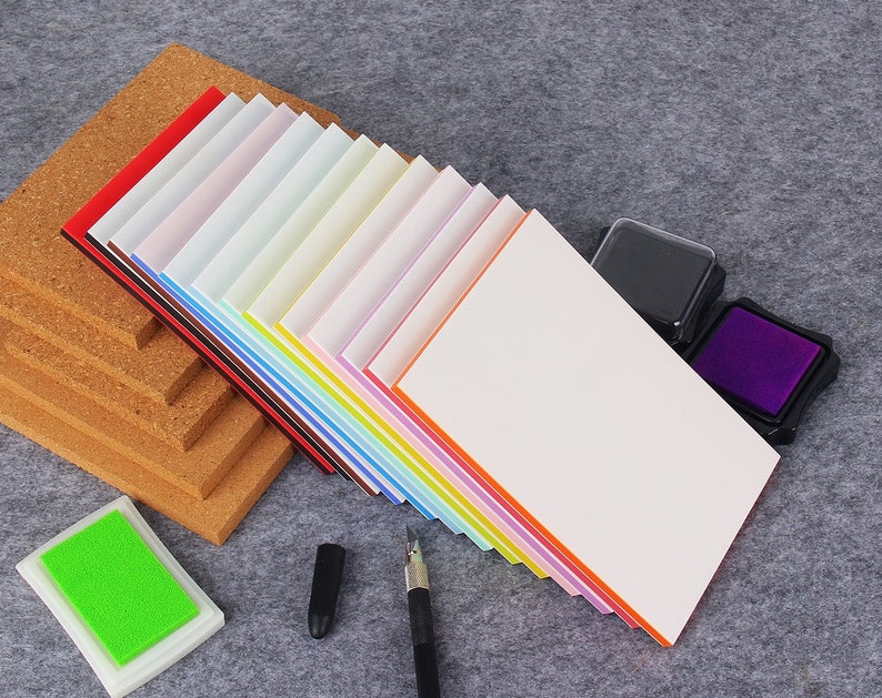 May include: A stack of colorful paper with a white top sheet, two ink pads, a pen, and a cork board. The paper has a variety of colors, including red, orange, yellow, green, blue, purple, and pink.