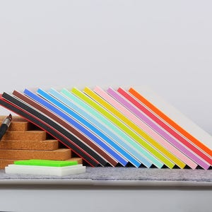 May include: A set of 15 colorful ink pads with white covers and black edges. The ink pads are arranged in a rainbow pattern, with red at the top and purple at the bottom. The ink pads are sitting on a cork surface.