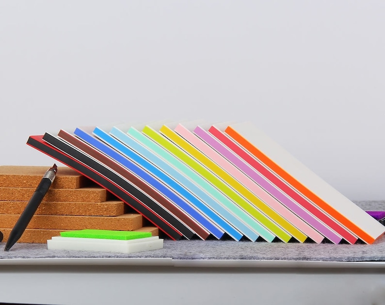 May include: A set of 15 colorful ink pads with white covers and black edges. The ink pads are arranged in a rainbow pattern, with red at the top and purple at the bottom. The ink pads are sitting on a cork surface.