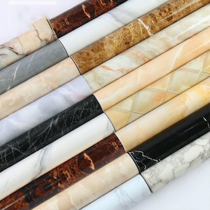 Puede incluir: Un surtido de rollos de papel de contacto con estampado de mármol en diferentes colores, incluyendo blanco, negro, marrón y beige. Los rollos están dispuestos en un patrón escalonado, con los extremos de los rollos visibles.
