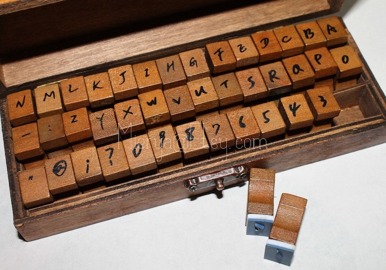 Pu&ograve; includere: Scatola di legno piena di timbri individuali di lettere e numeri. I timbri sono disposti in una disposizione simile a una tastiera. La scatola ha un fermo sul davanti. Due timbri aggiuntivi sono fuori dalla scatola.