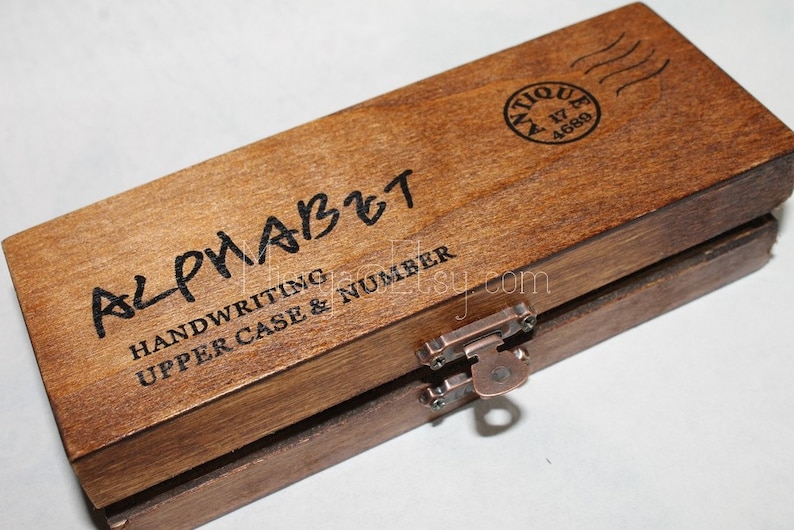 Pu&ograve; includere: Una scatola di legno con un chiavistello e un buco della serratura. La scatola &egrave; etichettata "Alphabet Handwriting Upper Case & Number". La parte superiore della scatola ha un timbro che dice "Antique 17 4689".