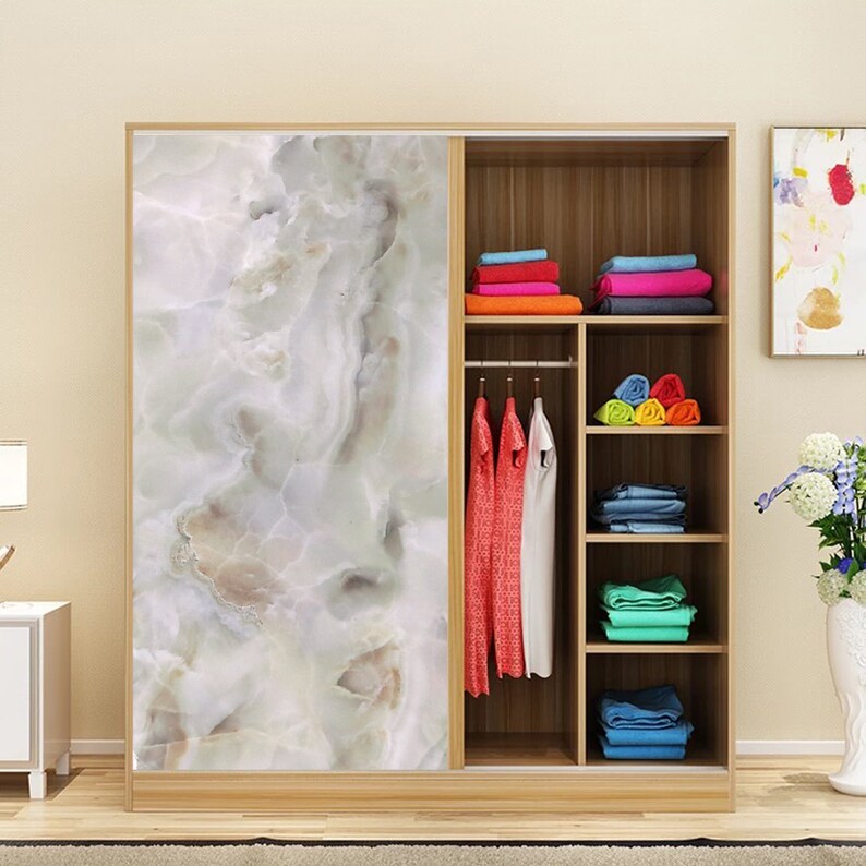 May include: A white wooden wardrobe with sliding doors. One door features a marble-like pattern, while the other door reveals an open interior with shelves and a hanging rod. The shelves are filled with folded clothes and the hanging rod has clothes hanging on it.
