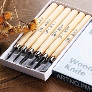 Puede incluir: Conjunto de seis herramientas de talla de madera con mangos de madera y cuchillas de metal. Las herramientas están en una caja blanca con las palabras "Wood Knife" y "ART.NO.PM1" impresas en la caja.