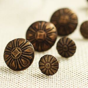 Puede incluir: Un conjunto de seis tachuelas de lat&oacute;n antiguo con un dise&ntilde;o floral decorativo. Las tachuelas est&aacute;n dispuestas en una fila sobre una superficie de tela blanca.