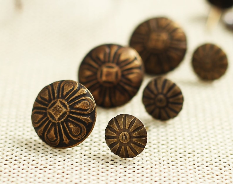 Puede incluir: Un conjunto de seis tachuelas de lat&oacute;n antiguo con un dise&ntilde;o floral decorativo. Las tachuelas est&aacute;n dispuestas en una fila sobre una superficie de tela blanca.