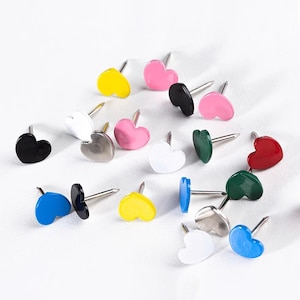 May include: A collection of heart-shaped push pins in various colors, including yellow, pink, black, white, silver, green, blue, and red. Each pin has a silver metal shaft. The pins are scattered on a white surface.
