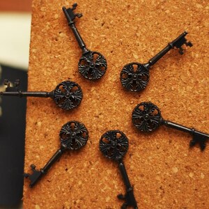 Op de afbeelding: Close-up van zes sierlijke, zwarte metalen skeletsleutels gerangschikt op een prikbord. Elke sleutel heeft een decoratieve, cirkelvormige boog en een gedetailleerde sleutelbaard. De sleutels zijn donker, matzwart en het prikbord biedt een warme, neutrale achtergrond.