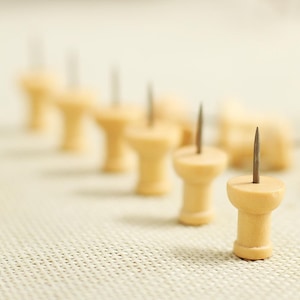 May include: A row of wooden push pins with metal points. The push pins are arranged in a line on a beige fabric surface.