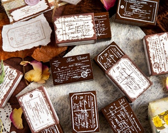 May include: A collection of vintage-style wooden stamps with handwritten text and designs. The stamps are brown with white lettering and are arranged on a brown surface with dried flowers and leaves.
