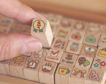 May include: A wooden box filled with small wooden stamps featuring various designs and words, including "check", "D-Day", "diet", and "P-S". A hand is holding a stamp with a red circle and the word "KITT" inside.