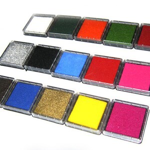 May include: A collection of ink pads in various colors, including white, green, red, and pink. Each pad is square and housed in a clear plastic case. The ink pads are arranged in rows, showcasing a range of vibrant hues suitable for crafts and stamping.